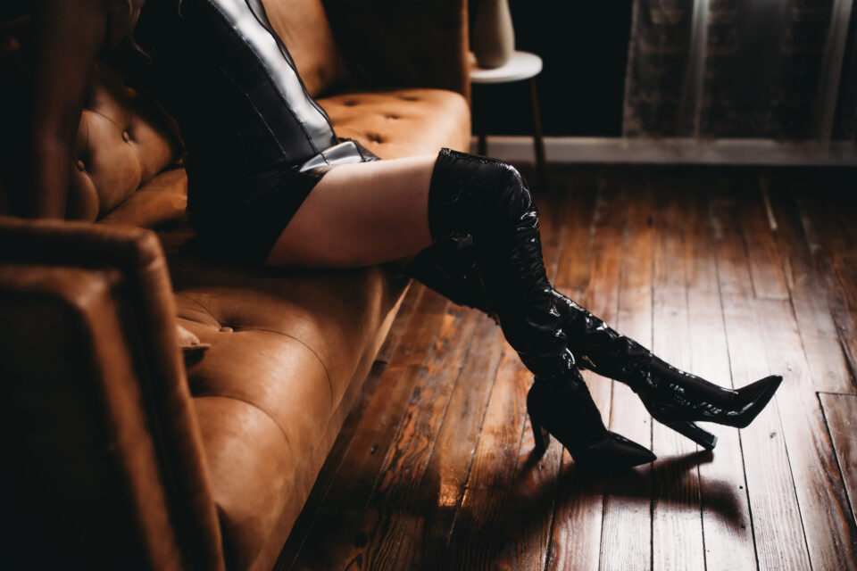 Dominatrix in latex and thigh-high boots leaning back on couch.