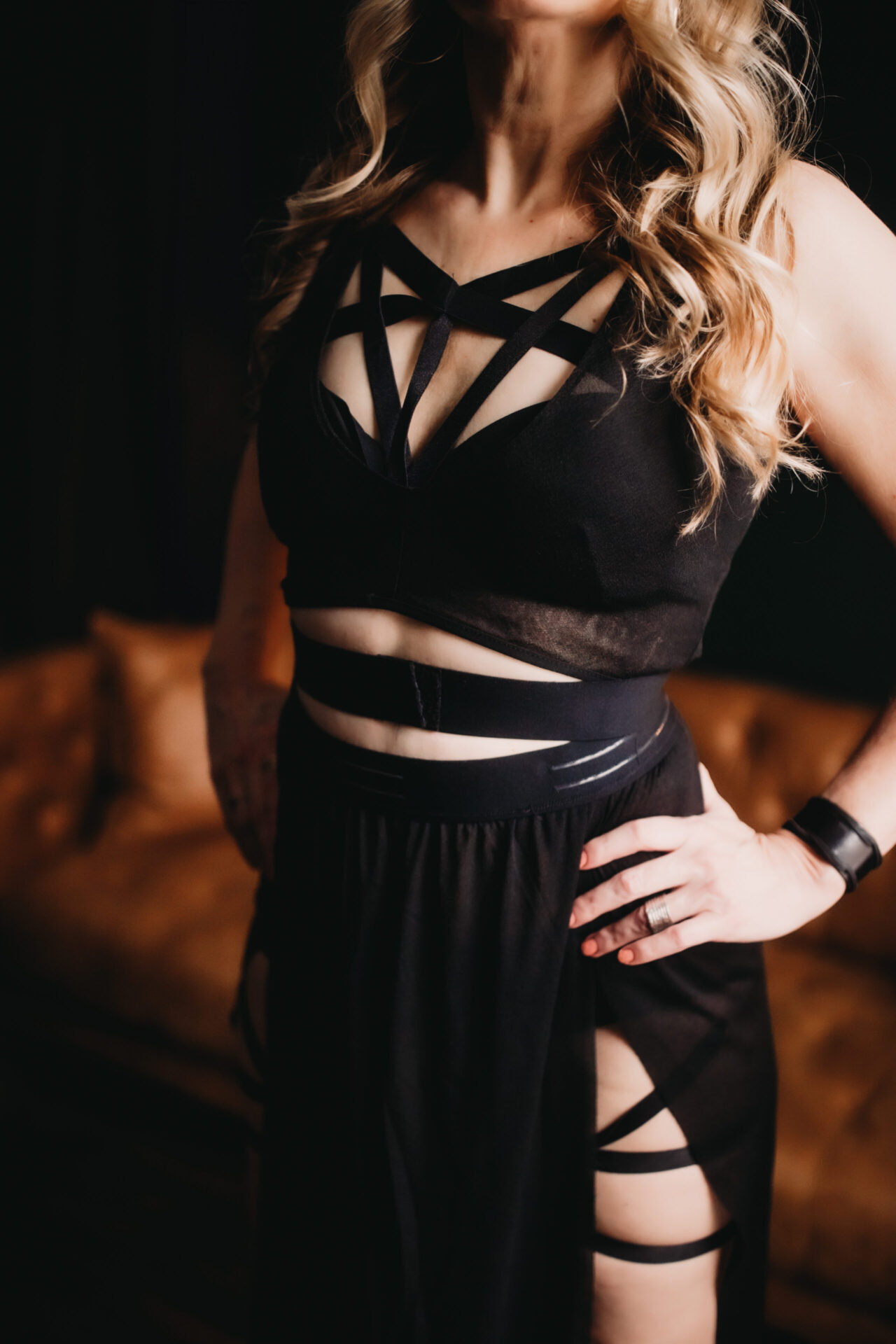 Dominatrix with hands on hips wearing black outfit with many straps over strips of tastefully exposed skin.