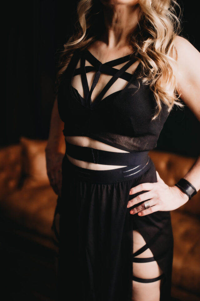 Dominatrix with hands on hips wearing black outfit with many straps over strips of tastefully exposed skin.