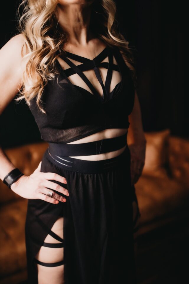 Dominatrix with hands on hips wearing black outfit with many straps over strips of tastefully exposed skin.